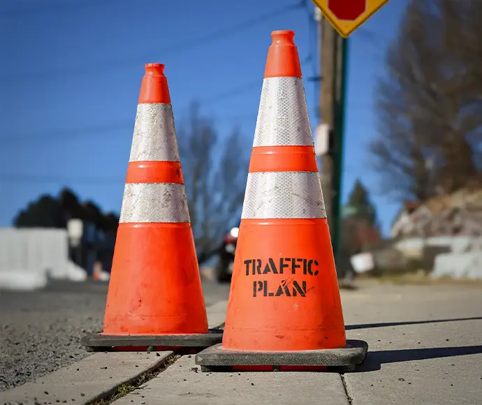Two orange traffic cones are placed on a sidewalk, with TRAFFIC PLAN stenciled in black on one cone. The background includes a street, a road sign, and leafless trees under a clear blue sky.