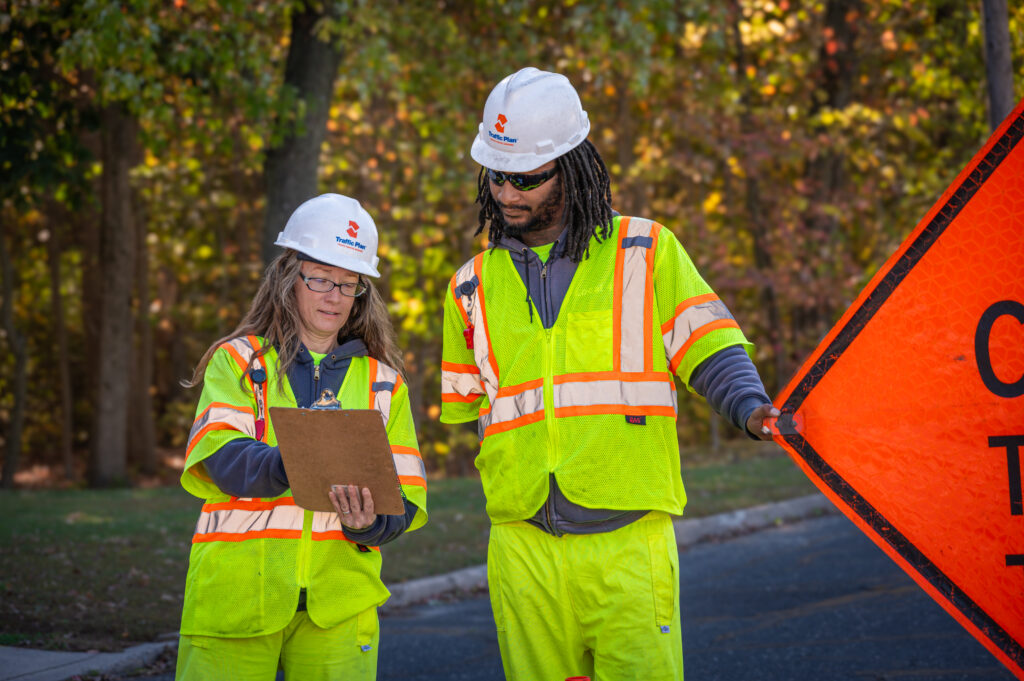 Two construction workers in reflective safety gear and helmets stand outdoors. One holds a clipboard and writes, while the other holds an orange roadwork sign. Trees and a road appear in the background.