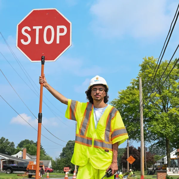 A construction worker in a neon safety vest and hard hat holds up a large stop sign on a suburban street, with trees, power lines, and houses in the background on a clear day.