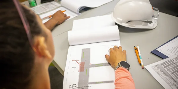 A person sits at a desk with construction plans, a pencil, and documents. A white hard hat, drink bottle, and coffee cup are on the table. The person is reviewing a diagram with labeled measurements.