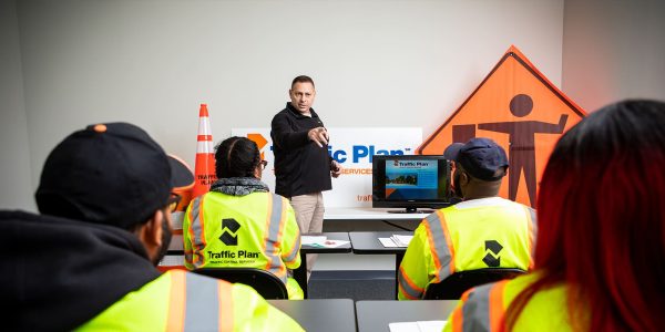A man stands at the front of a classroom pointing while instructing four people in safety vests. Traffic safety signs, a cone, and a presentation screen are visible in the background.