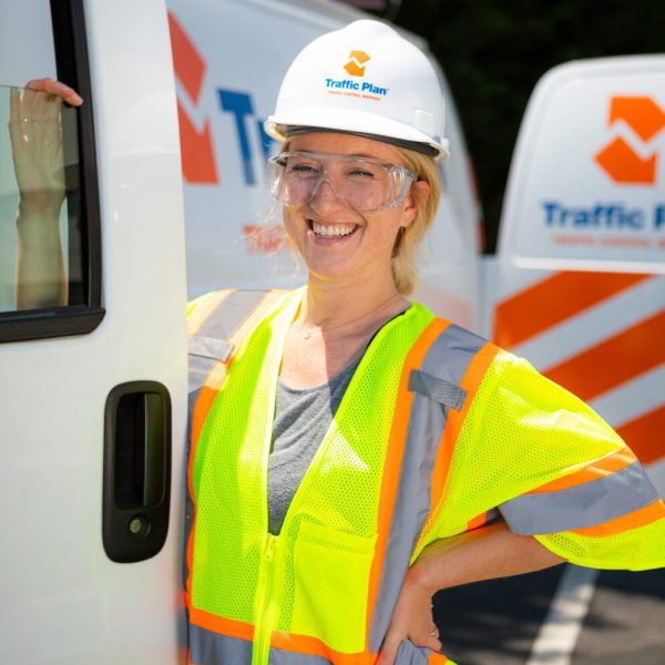 A smiling construction worker wearing a hard hat, safety glasses, and a neon yellow safety vest stands by a white van with Traffic Plan logos and orange stripes visible in the background.