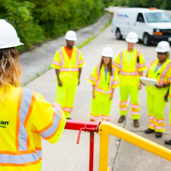 A group of five construction workers in neon safety gear listens to a supervisor standing on a platform. A white van and traffic cones are visible on the road, with trees lining the background.