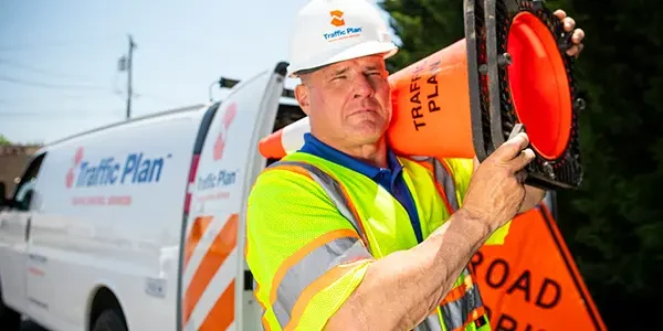 A construction worker in a reflective vest and hard hat carries an orange traffic cone over his shoulder, standing near a van and a Road Work Ahead sign on a sunny day.