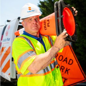 A construction worker in a reflective vest and hard hat carries an orange traffic cone on his shoulder, standing near a Road Work Ahead sign and a work vehicle.