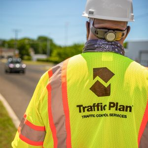 A person wearing a hard hat and a neon safety vest labeled Traffic Plan Traffic Control Services stands beside a road with a blurred car in the background.