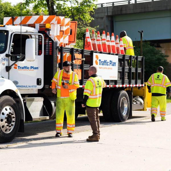 Four road workers in high-visibility clothing stand near a work truck loaded with traffic cones on the side of a road, with an overpass and greenery in the background.