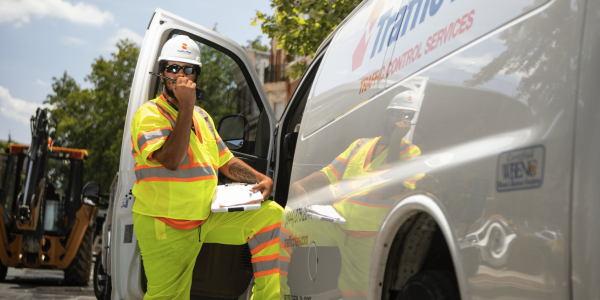 A construction worker in a neon safety uniform and hard hat stands by a white van with Traffic Plan signage, holding a clipboard and talking on a two-way radio. Construction equipment and trees are in the background.